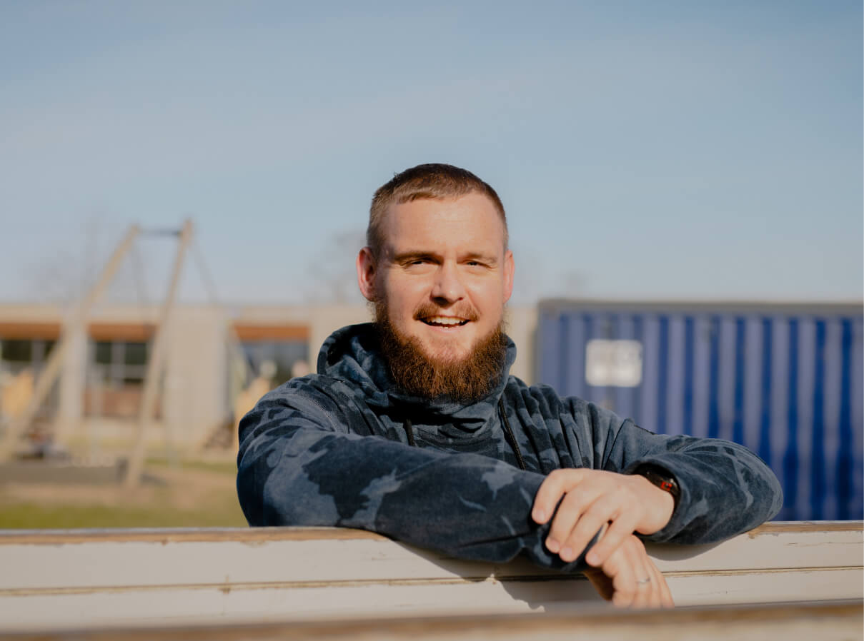 Niklas, mandlig pædagog, står udenfor og smiler til kameraet med hænderne hvilende på et hegn