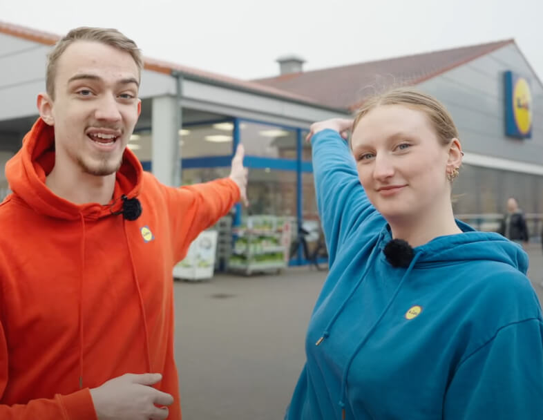 To butikselever fra Lidl står foran facaden på en Lidl-butik og ser glade ud