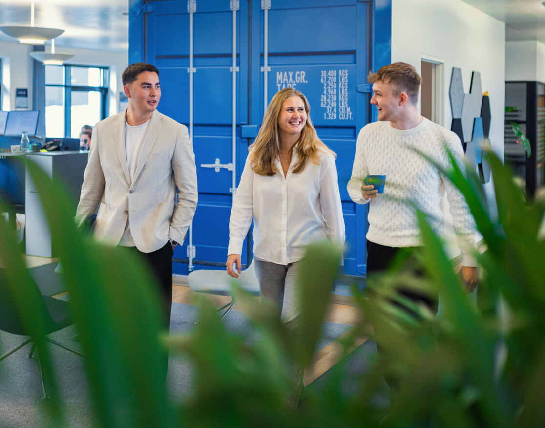 Tre smilende elever kommer gående ned af gang i kontormiljø ved Kuehne + Nagel 