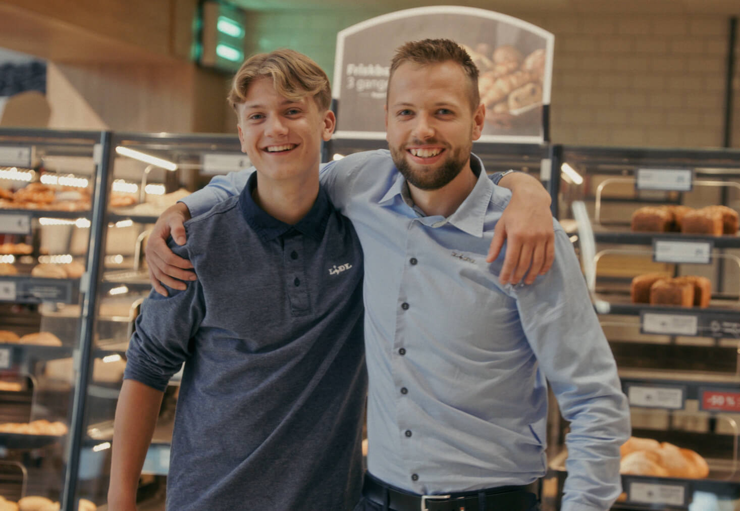 To Lidl-medarbejdere står med armen om hinanden og smiler foran bake off-afdelingen i Lidl