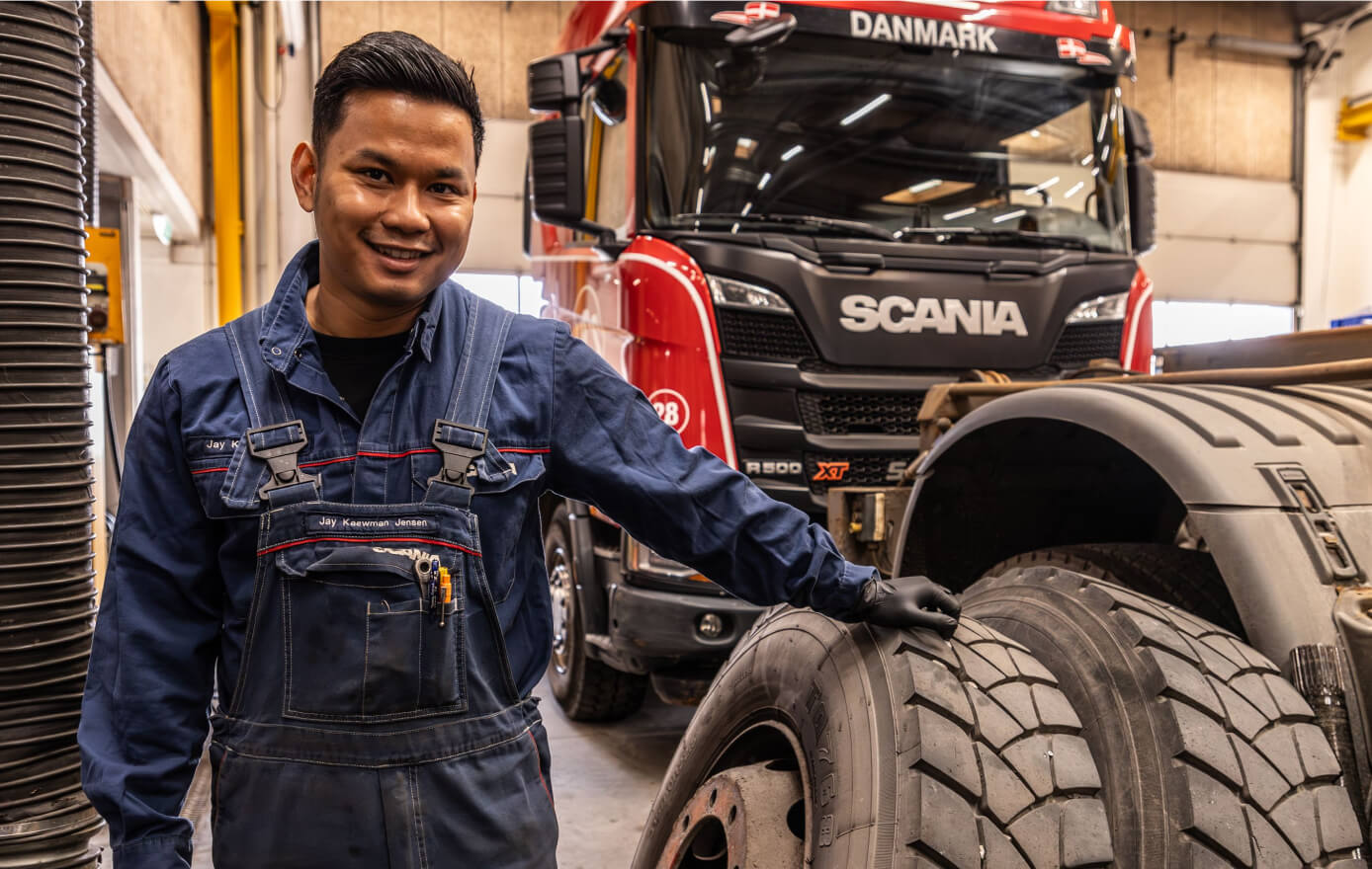 Mandlig mekaniker poserer smilende til kameraet, mens han læner sig op af et stort dæk